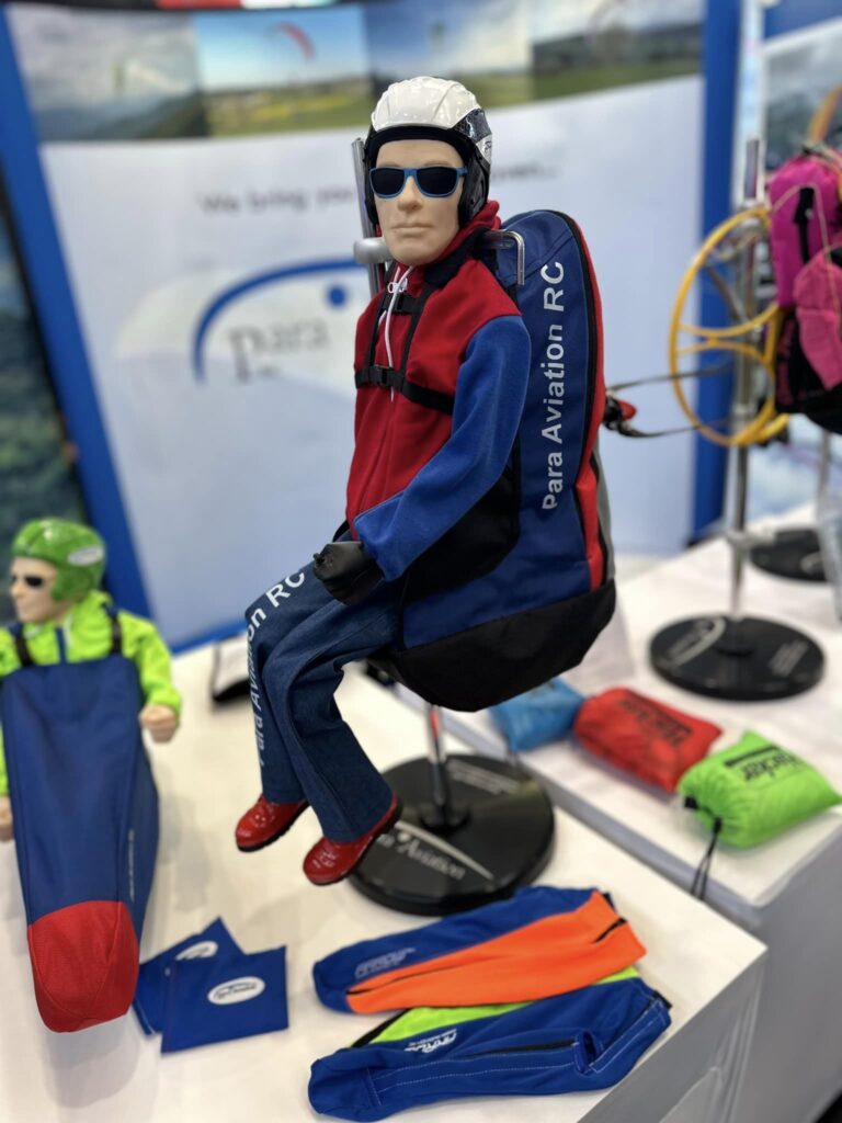 Die Para Aviation RC-Pilotenfigur „NOAH“ sitzt zusammen mit dem „NOAH Sitzgurtzeug“ auf einem Tisch an einem Messestand.