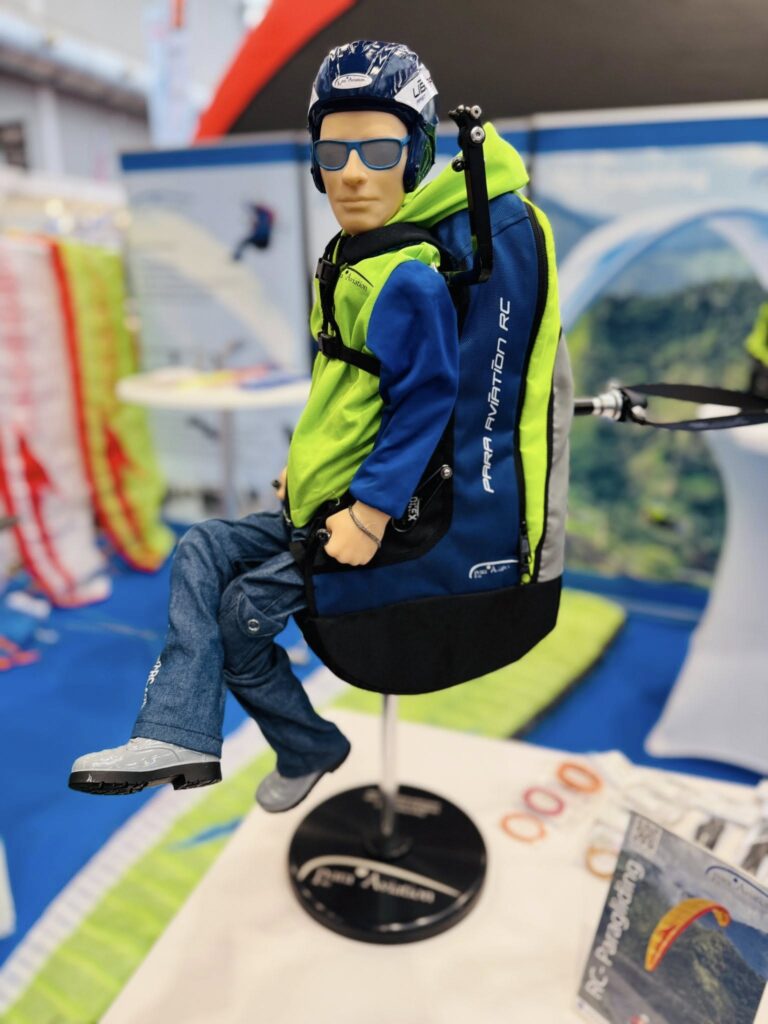 Die Para Aviation RC-Pilotenfigur „NOAH ST“ sitzt zusammen mit dem „Sitzgurtzeug Libra ST“ auf einem Tisch an einem Messestand.